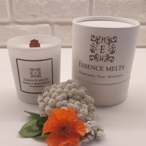 White glass candle labeled Verbena & Blood Orange, placed on a rustic wooden surface with a blurred natural background. Spa Candles