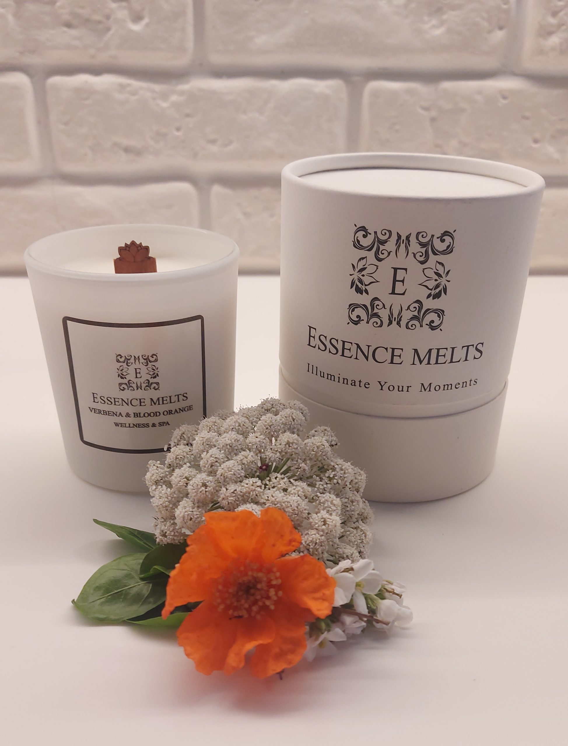 White glass candle labeled Verbena & Blood Orange, placed on a rustic wooden surface with a blurred natural background. Spa Candles