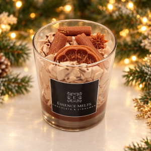 Chocolate and cinnamon candle with whipped wax and spice-shaped wax decorations in a glass, surrounded by Christmas greenery and golden lights.