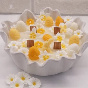 Easter-themed candle with yellow and white wax flowers, eggs, and bunnies in a scalloped white ceramic bowl., easter scented candle