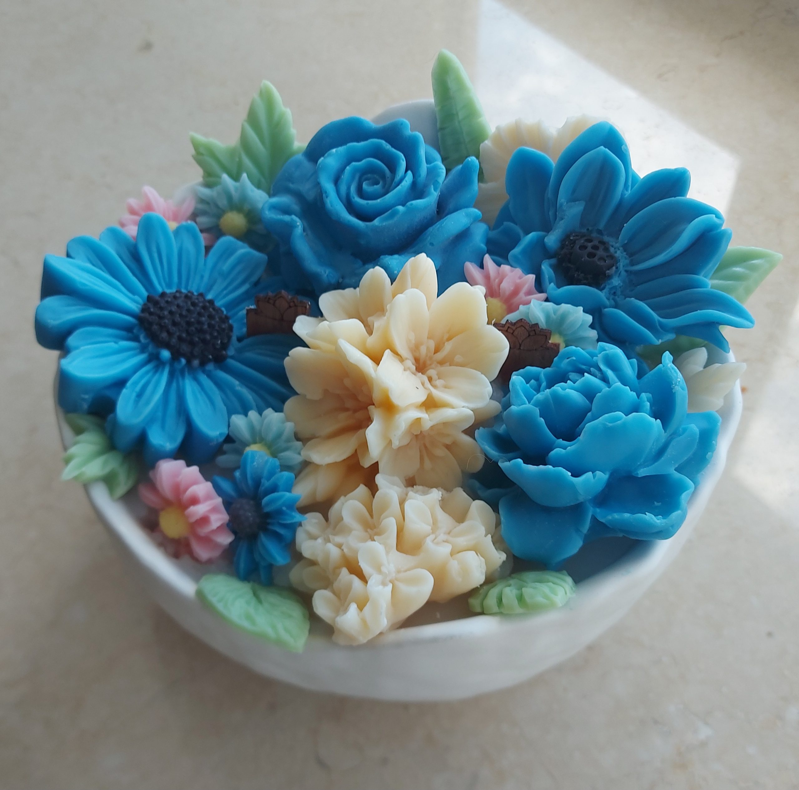 Jasmin Bloom Candle featuring a white bowl adorned with blue, pink and white flowers of wax scented with Jasmin. in between the flowers are the green was leaves.