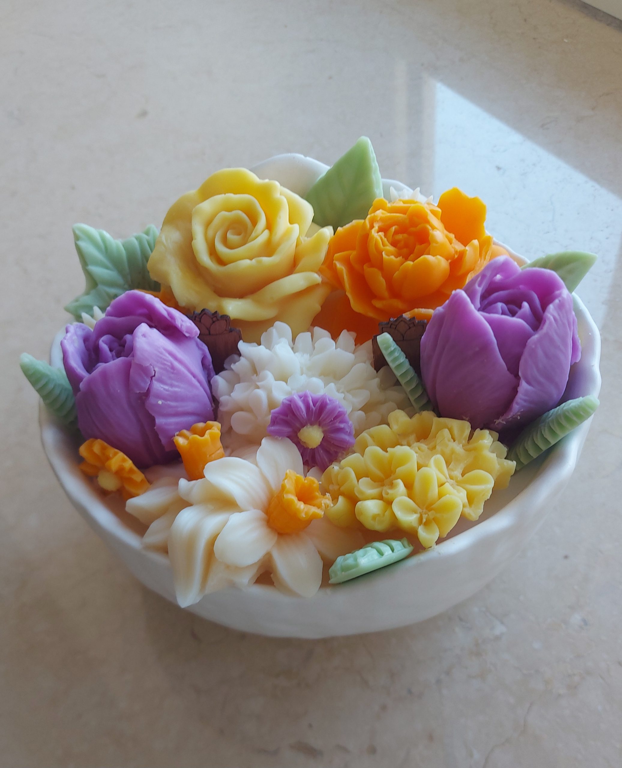 Hand-poured Monoi Bloom candle in a white ceramic bowl, decorated with vibrant wax flowers in yellow, white, lilac, and orange tones, with green wax leaves.