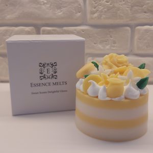 A Candle looking like a lemon meringue cake in white and yellow colors, topped with a yellow rose and green leaves and mini lemons of wax.