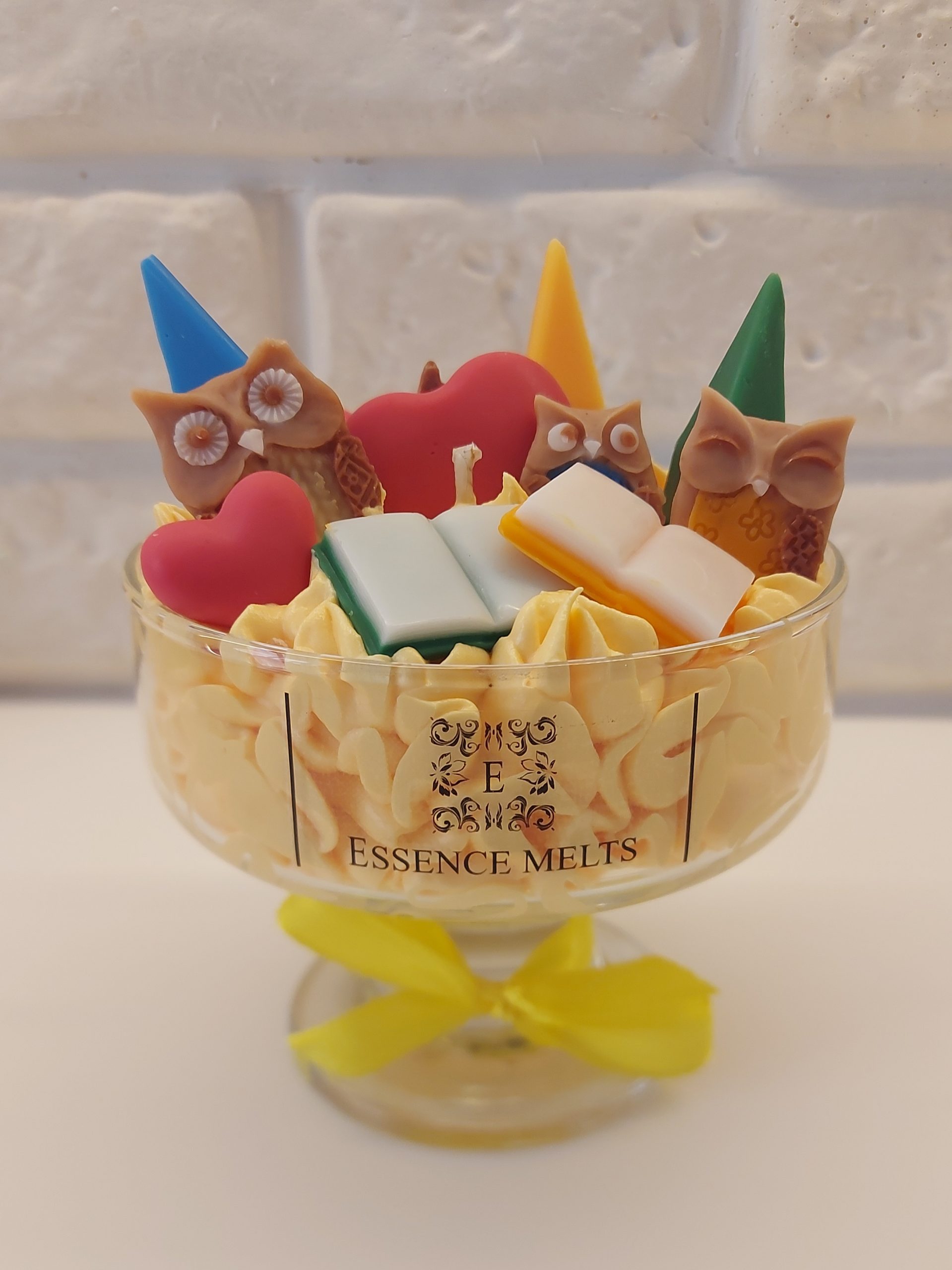 Colorful Best Teacher candle in a glass container with a yellow ribbon. red, blue and yellow wax crayons, 3 owls of wax and 2 colorful wax books, red apple and red hearts on top of a piped yellow wax.