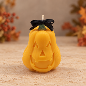 Orange jack-o’-lantern candle with a green stem and black satin bow, displayed on a stone surface with autumn leaves behind it.