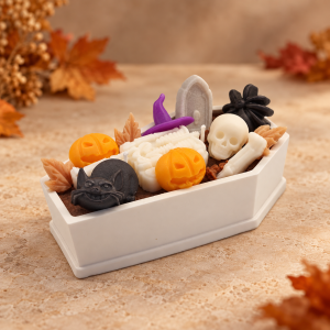 keleton candle in a white coffin-shaped vessel, decorated with a skull, bones, orange pumpkins, black cat and purple witch hat.